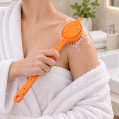 A person in a white robe uses an orange silicone back scrubber with soap suds on their shoulder in a bathroom.