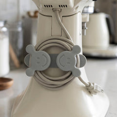 A gray silicone cord organizer with two smiling faces, holding a coiled light gray appliance cord on a white stand mixer.