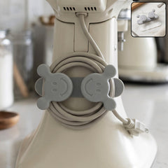 A gray silicone cord organizer with two smiling faces, holding a coiled light gray appliance cord on a white stand â€“ view 3