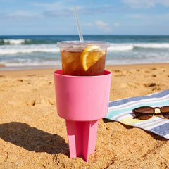 A pink plastic cup holder with an iced tea drink on a sandy beach, with a striped towel and sunglasses nearby.