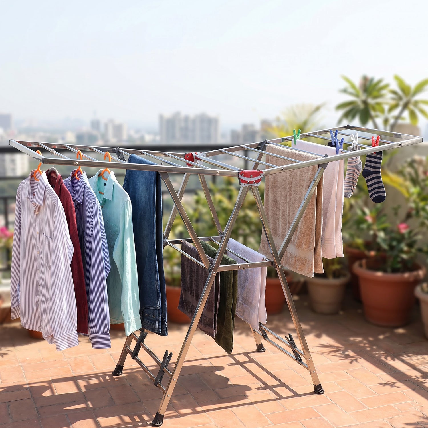 A silver clothes drying rack holding various garments, positioned on a sunny outdoor balcony with potted plants.