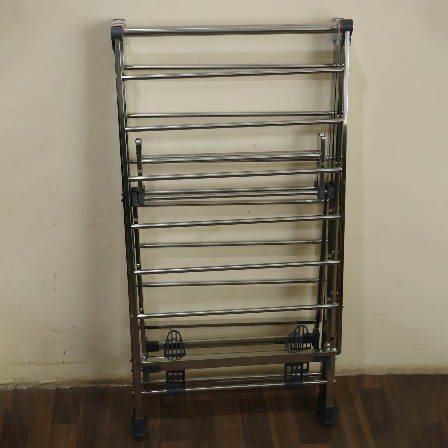 A silver metal clothes drying rack folded flat against a light brown wall, resting on a dark wooden floor.