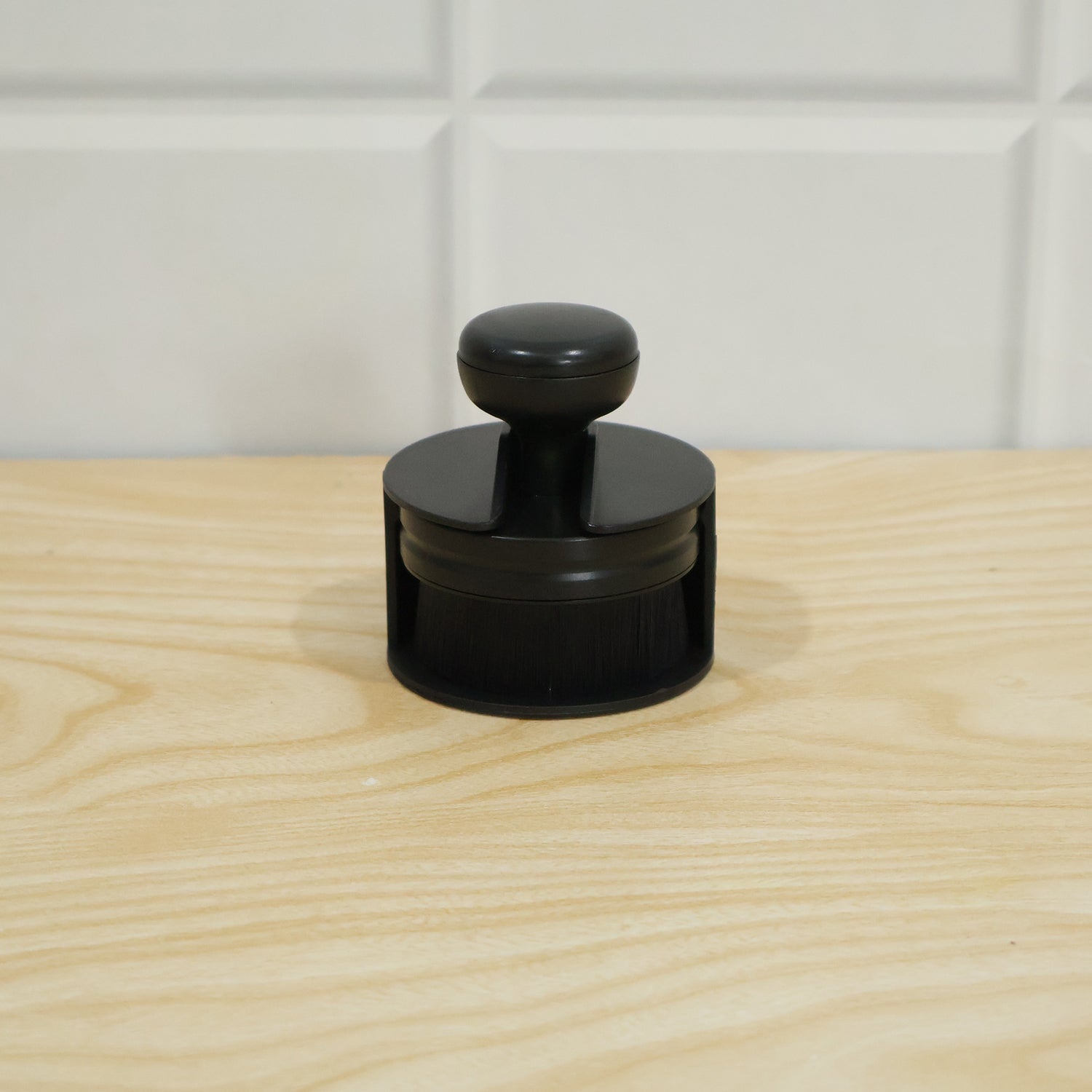 The black detailing brush is placed inside its cylindrical base on a light wooden surface, with a white tiled wall behind.