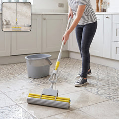 A person uses a yellow and grey sponge mop on a wet tiled kitchen floor, with a grey bucket nearby.