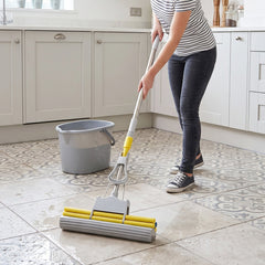 A person operates a yellow and grey sponge mop on a wet tiled kitchen floor, beside a grey bucket.