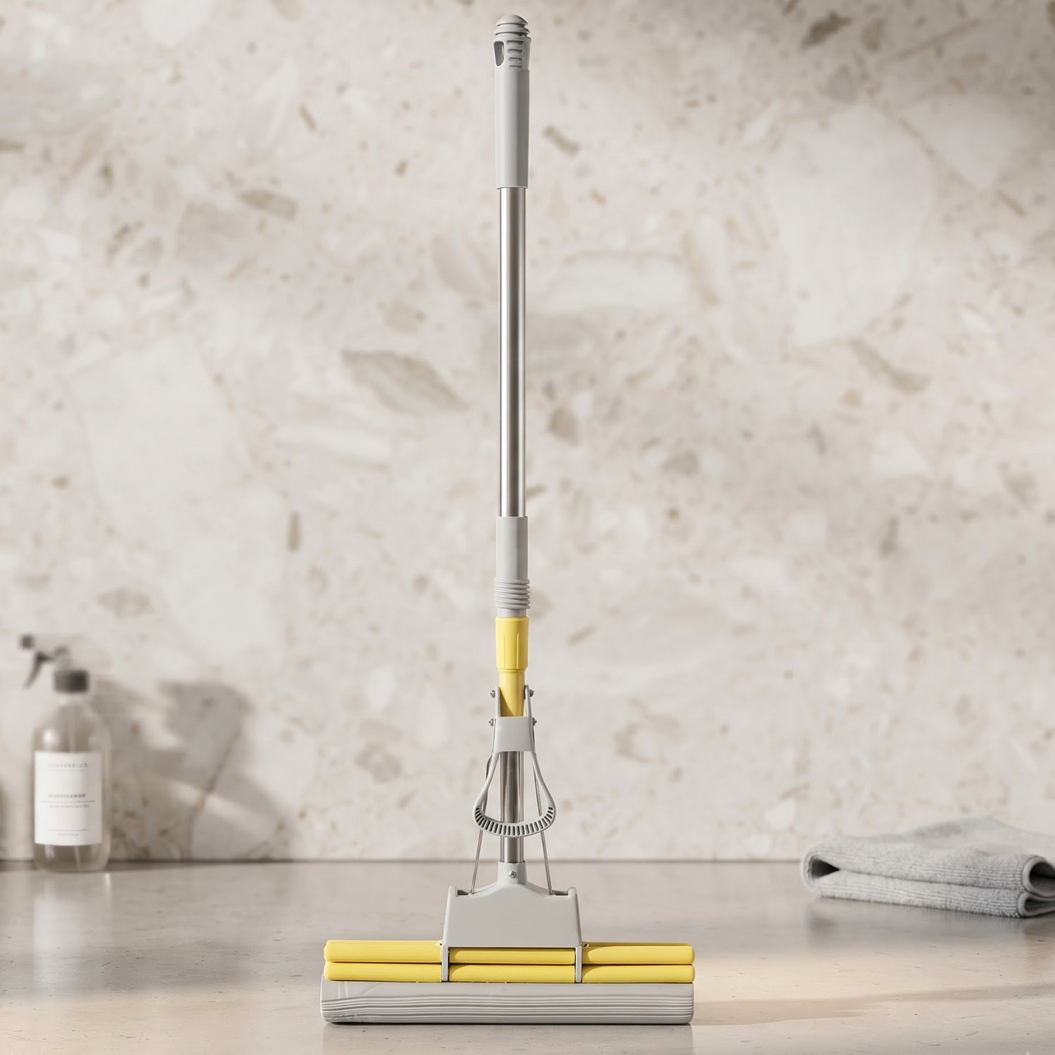 A yellow and grey sponge mop stands upright on a kitchen counter, with a spray bottle and towel nearby.