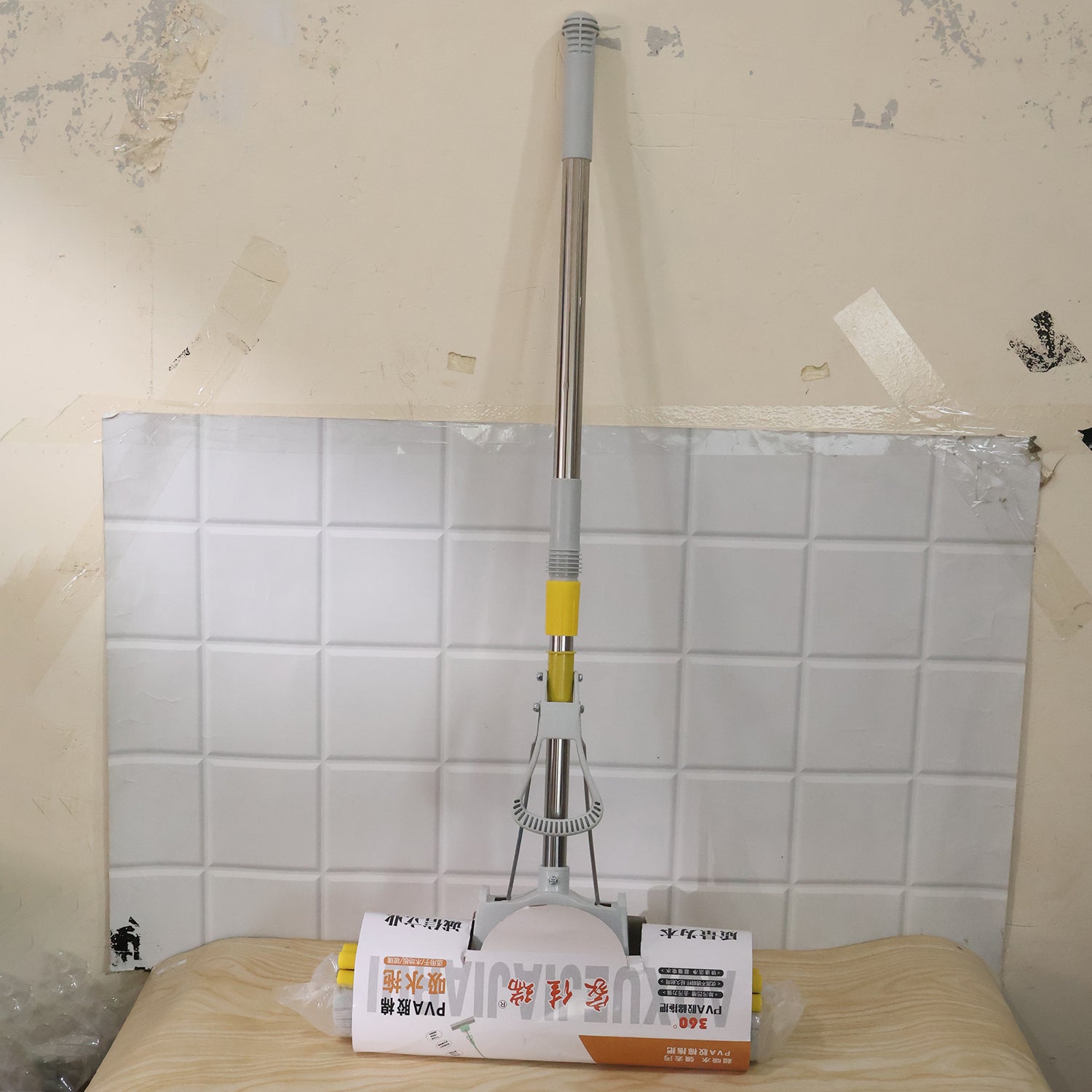 A yellow and grey sponge mop, still in its plastic packaging, rests on a wooden surface against a tiled wall.