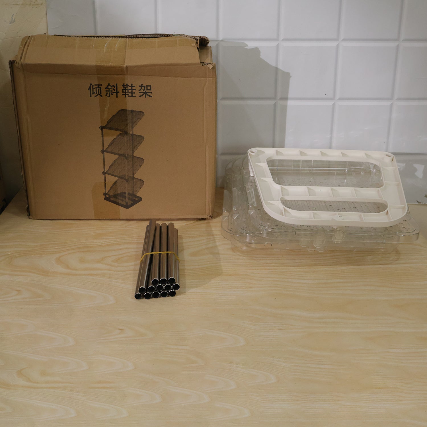 Disassembled shoe rack components, including a cardboard box, metal poles, and clear plastic shelves, on a wooden surface.