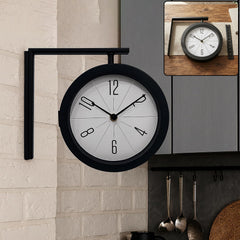 A black single-faced clock with a white background and clear numbers, mounted on a white brick wall.