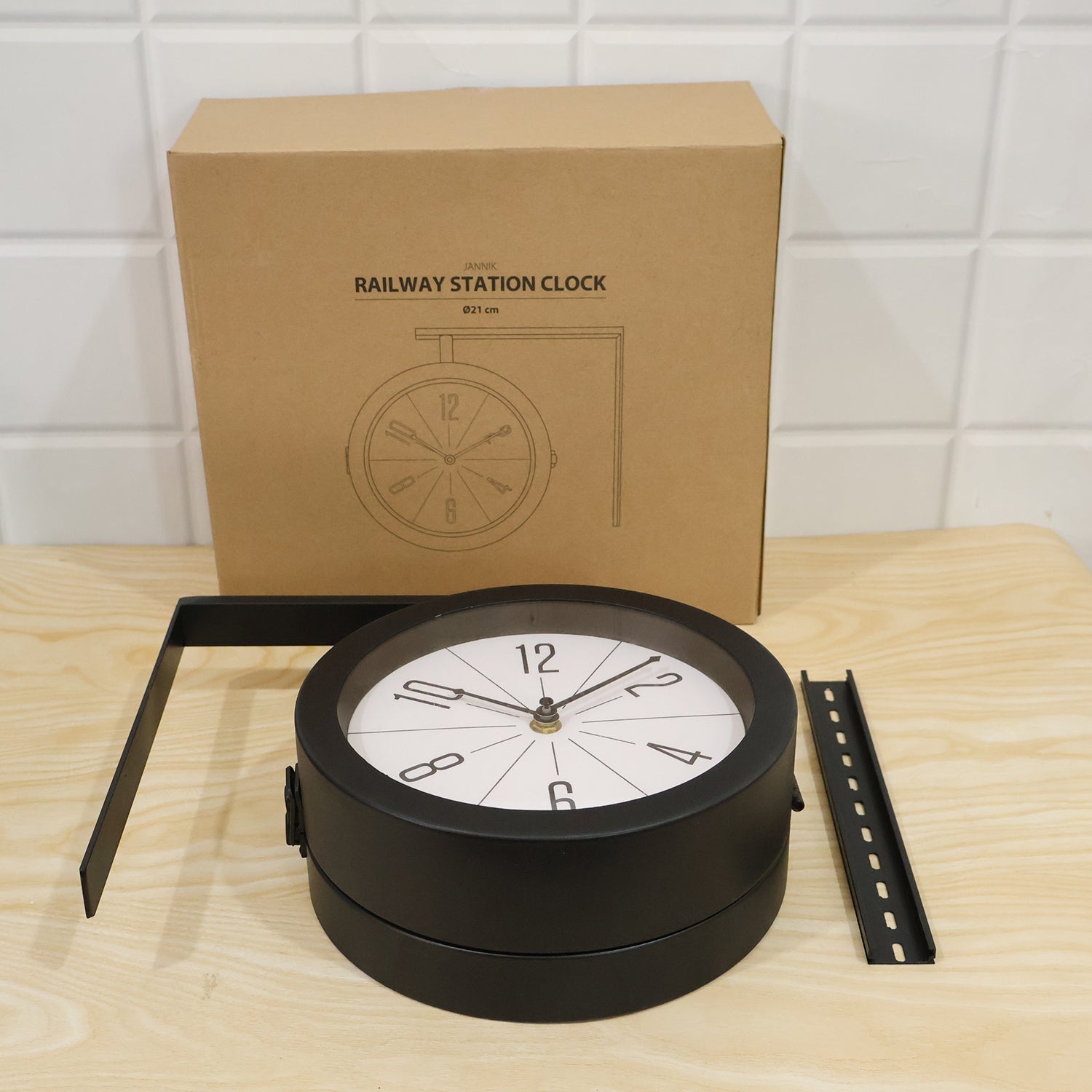 The black double-sided clock and its mounting bracket are displayed on a light wooden surface next to a brown cardboard box