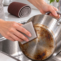 Hands scrubbing a dirty metal pot with a small gray and brown abrasive sponge in a kitchen sink.