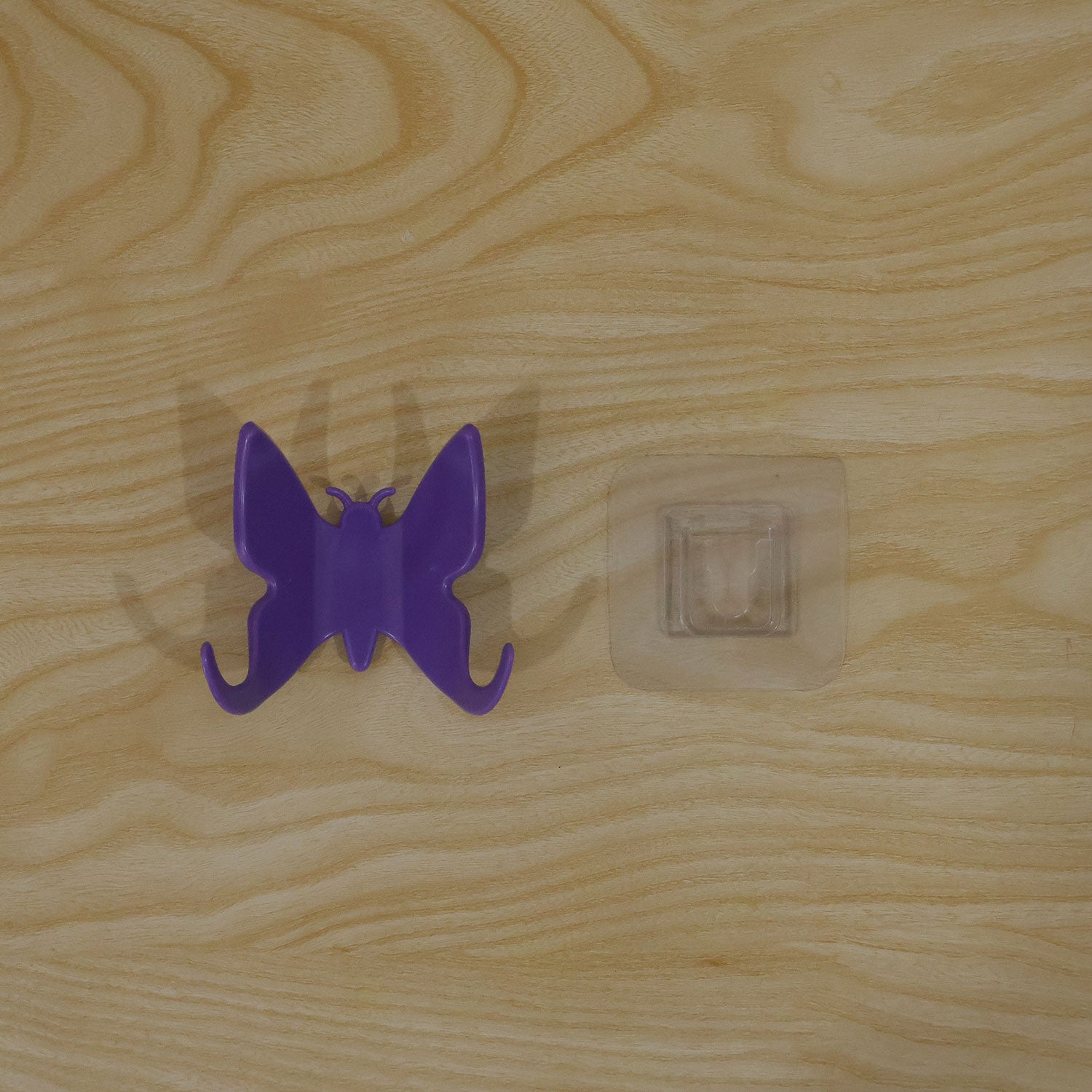 A purple butterfly hook and its clear adhesive mount are placed side-by-side on a light wooden surface.