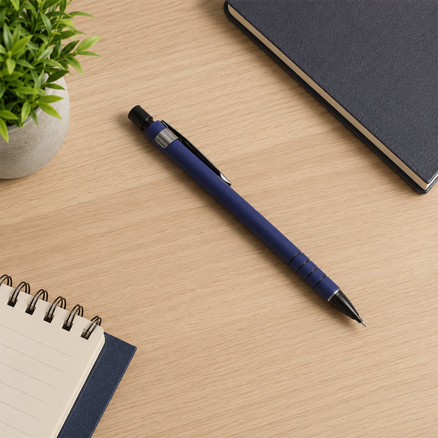 A blue mechanical pencil lies on a light wooden desk next to a green plant, a dark blue notebook, and a spiral notepad.
