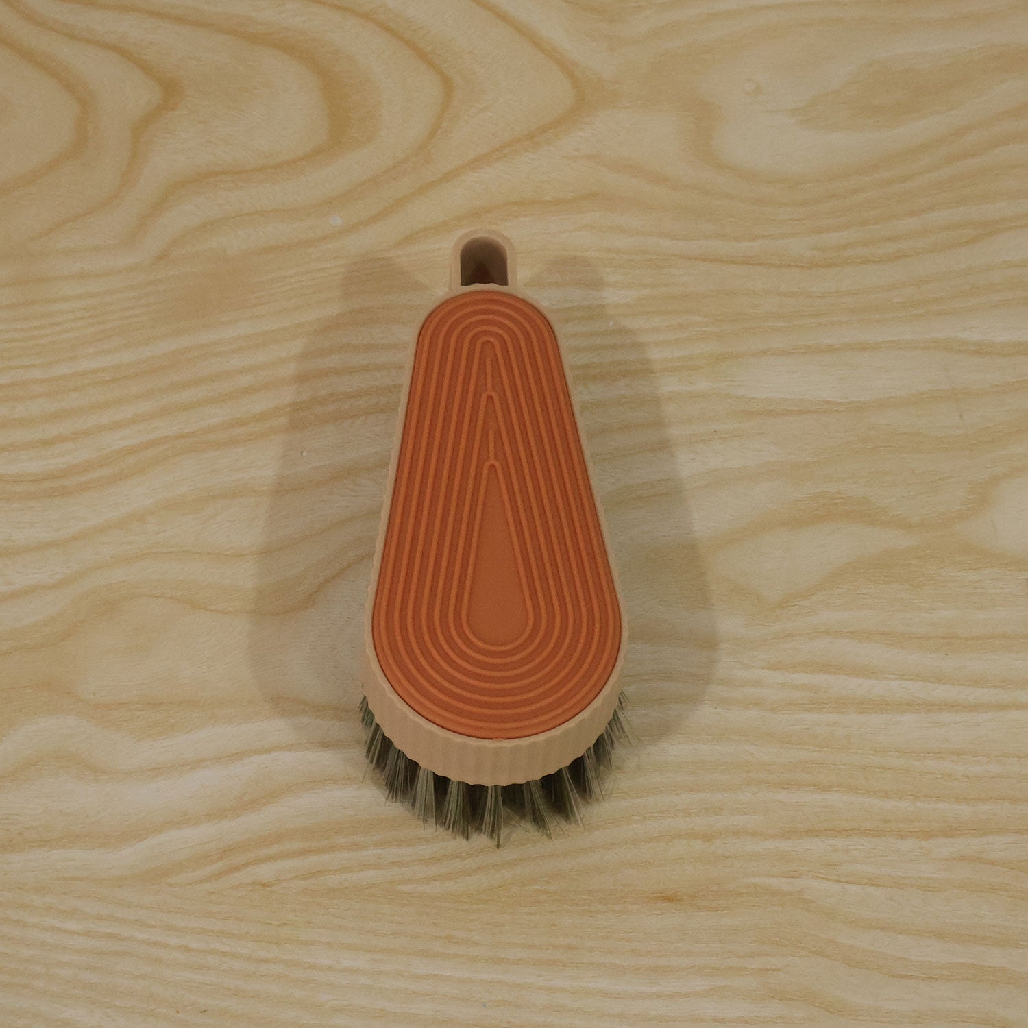 An oval-shaped beige brush with an orange textured top and green bristles is centered on a light wooden surface.