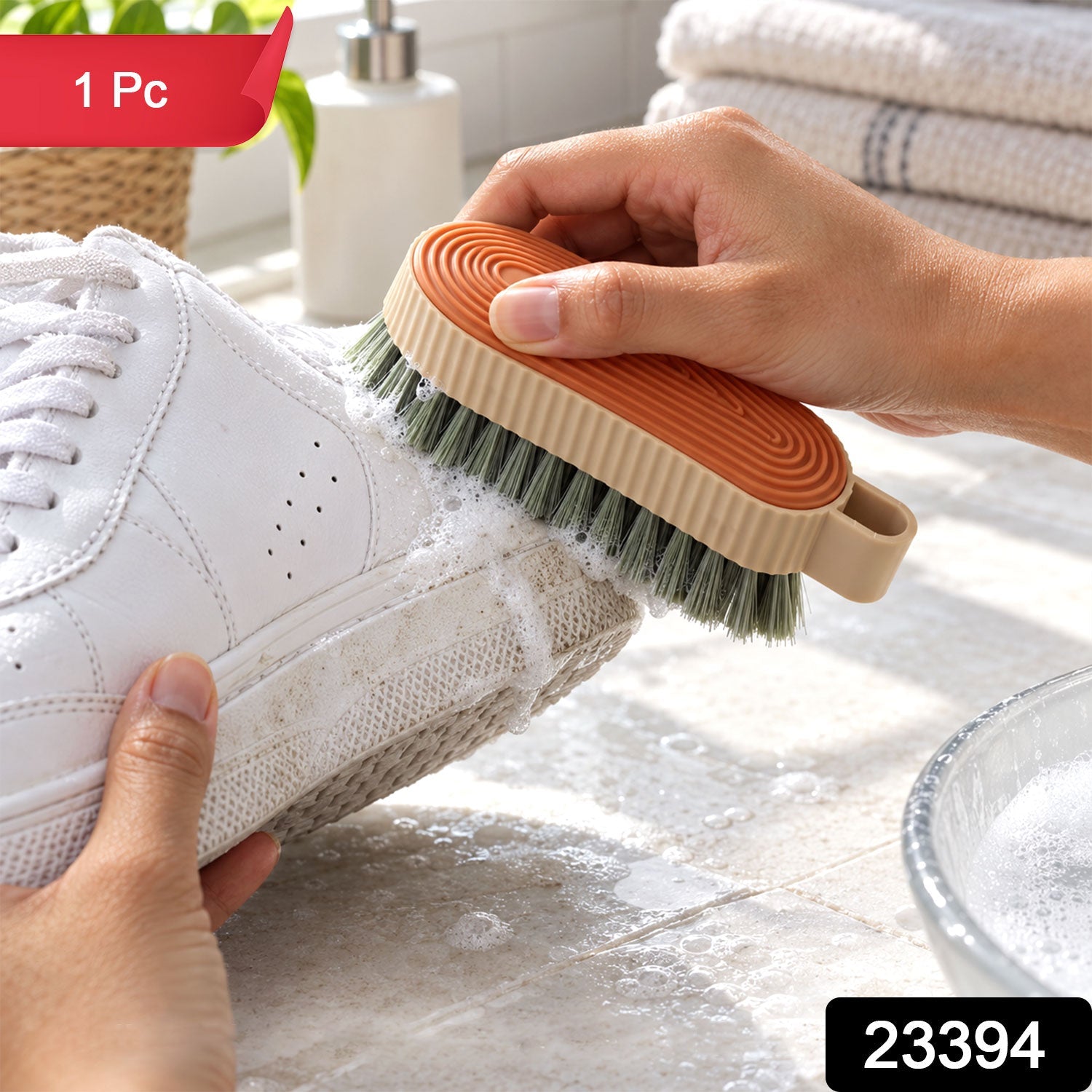 Hands clean a white shoe with a beige and orange brush, generating suds, with a soap dispenser and towels in the background.