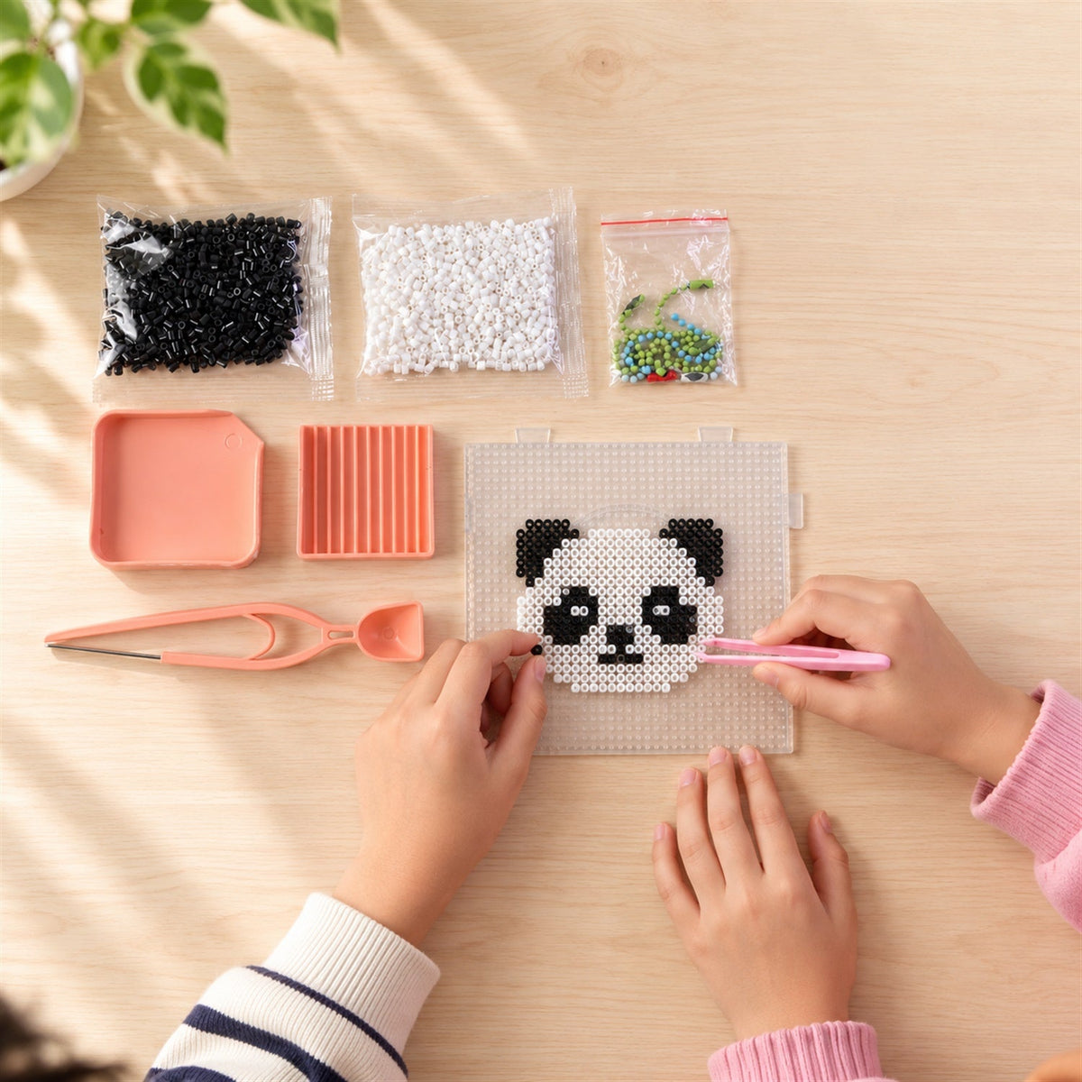 Overhead view of hands using pink tweezers to place black and white fuse beads on a clear pegboard, forming a panda design.