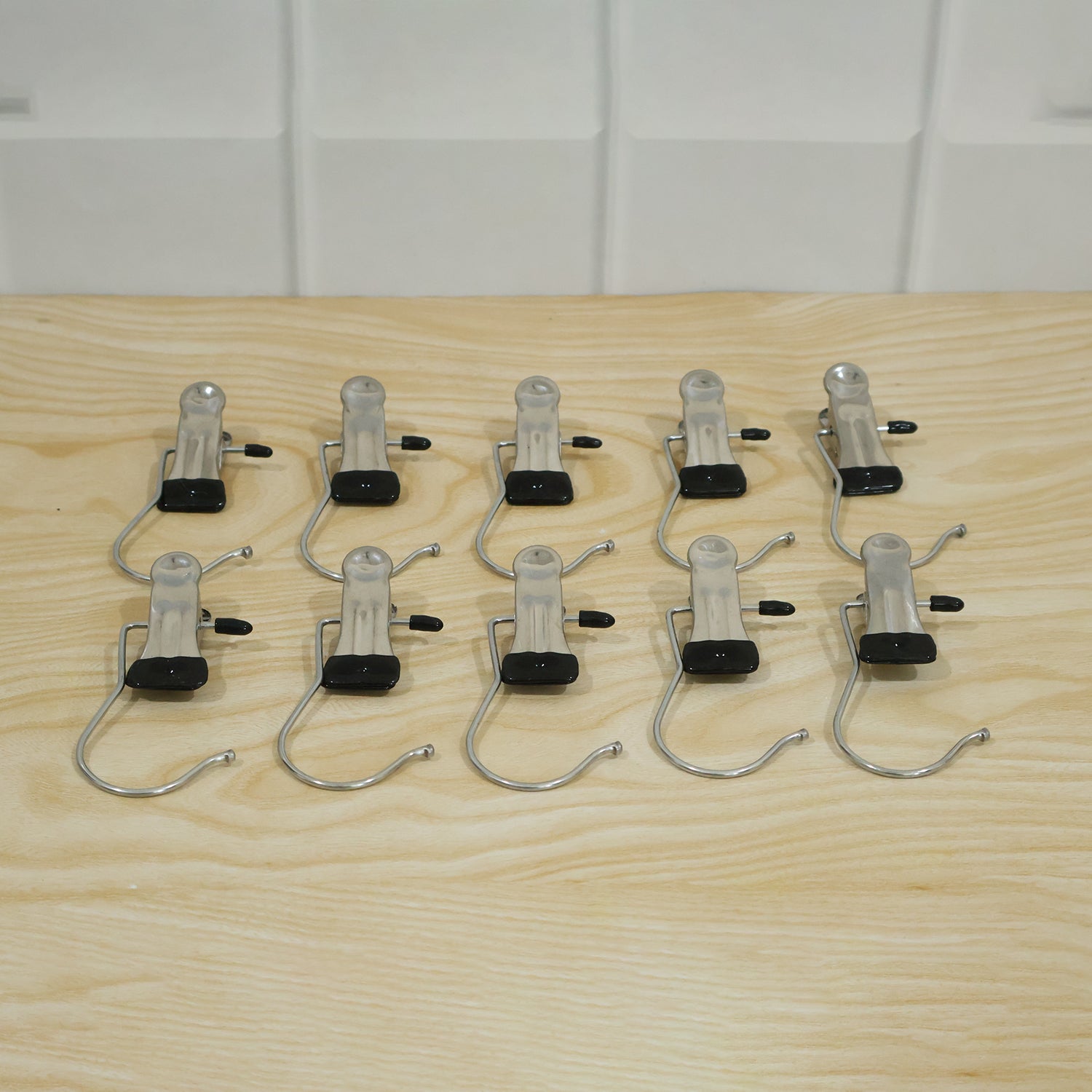 Ten metal clip hangers with black rubber tips and S-shaped hooks are arranged in two rows on a light wooden surface.