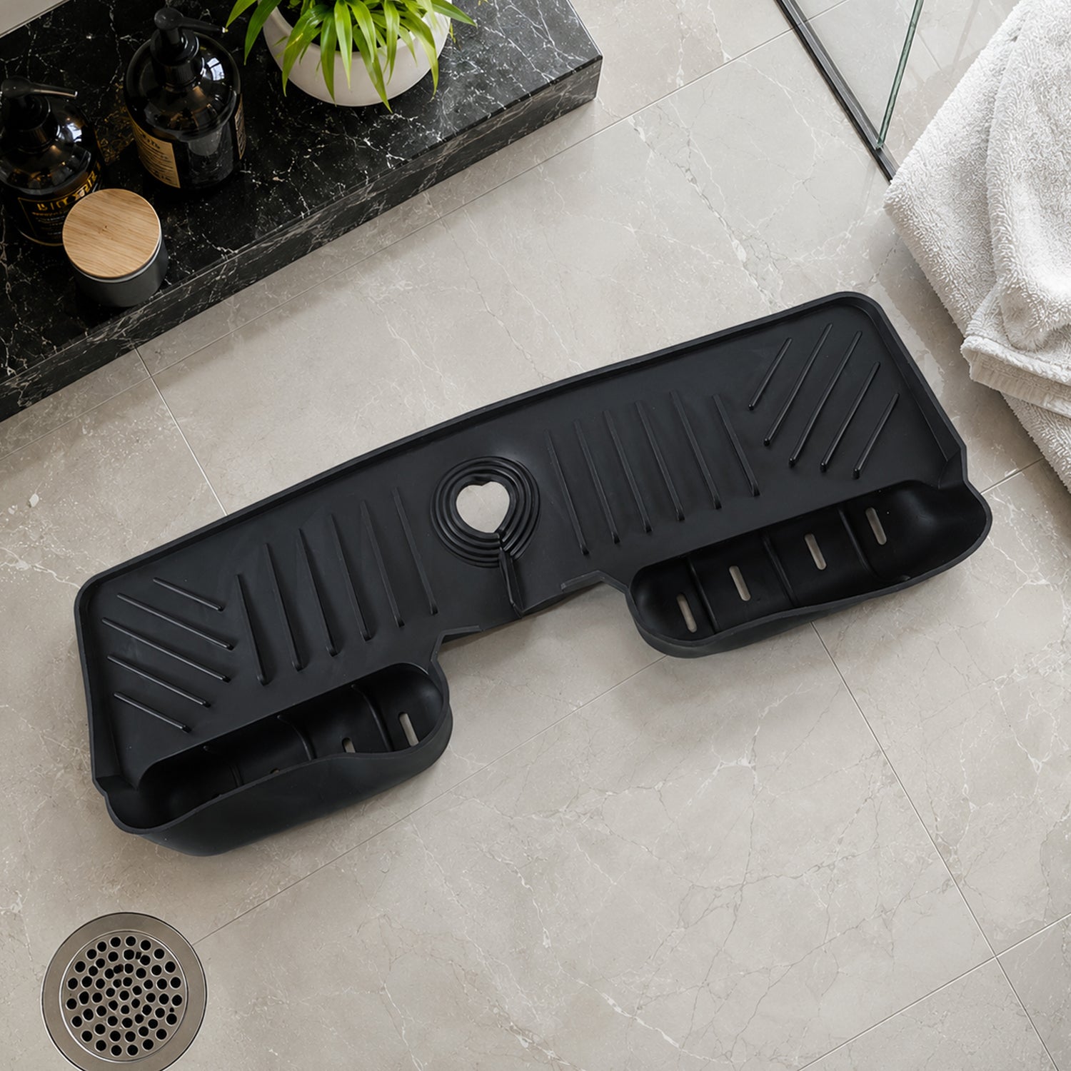 A black silicone faucet splash guard with drainage grooves and two side compartments, lying on a light grey tiled floor next