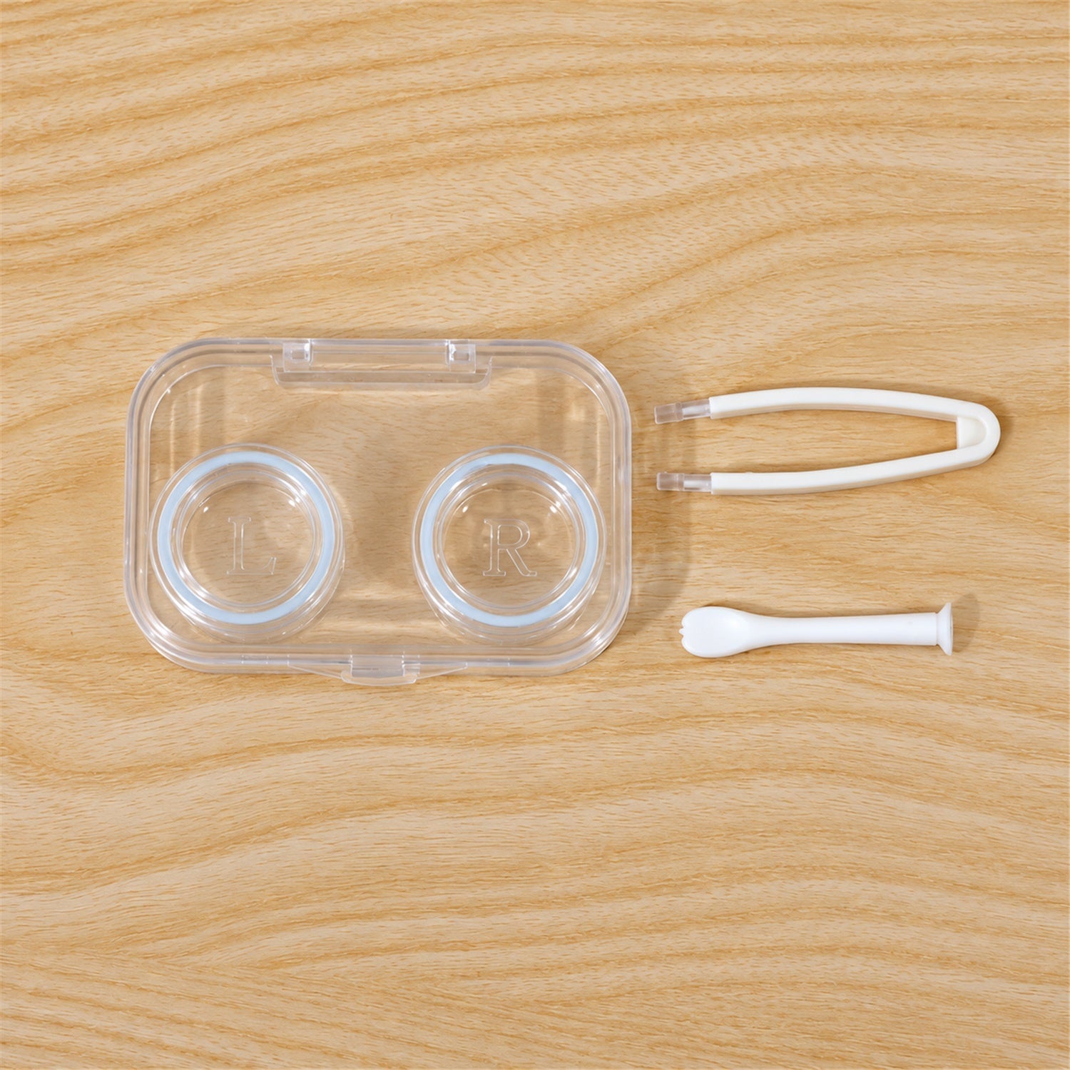 An open clear contact lens case with two compartments, white tweezers, and an applicator arranged on a wooden table.