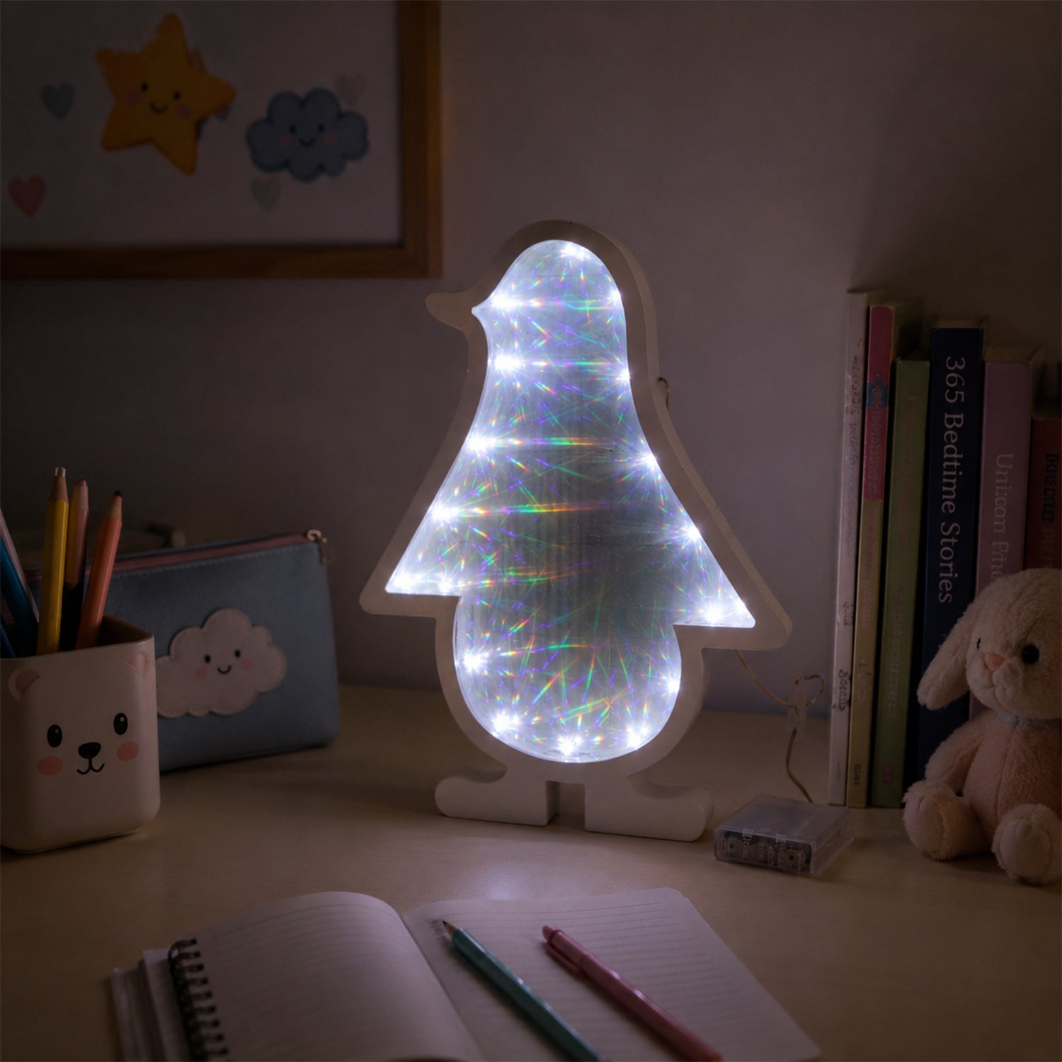 White penguin-shaped infinity mirror light glowing on a child's desk, surrounded by school supplies and a notebook.