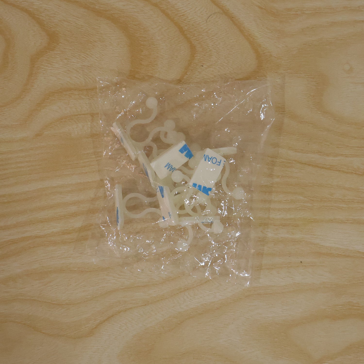 Several white plastic self-adhesive plant clips are visible inside a clear plastic bag on a light wooden surface.