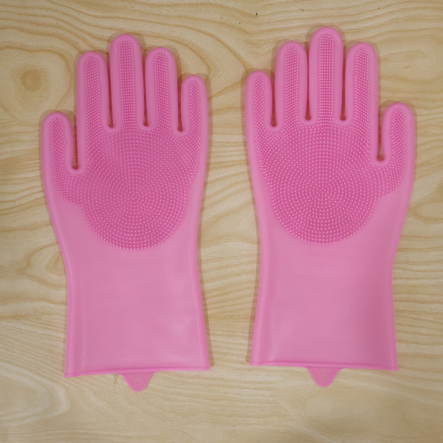 A pair of pink silicone scrubbing gloves with textured palms laid flat on a light wooden surface.