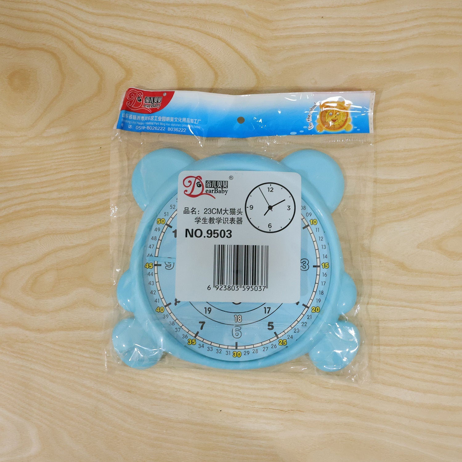 A light blue bear-shaped learning clock is sealed in clear plastic packaging with a white label on a wooden surface.