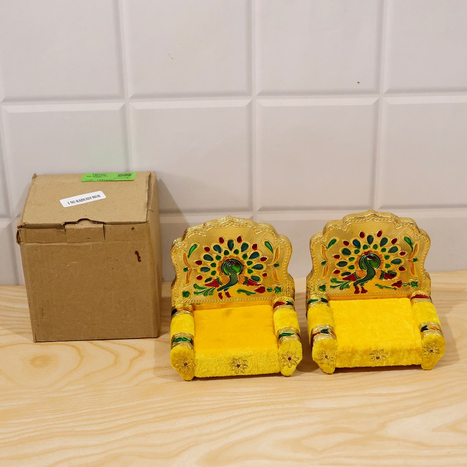 Two yellow singhasans with golden peacock backrests sit next to a small brown cardboard box on a light wooden table.
