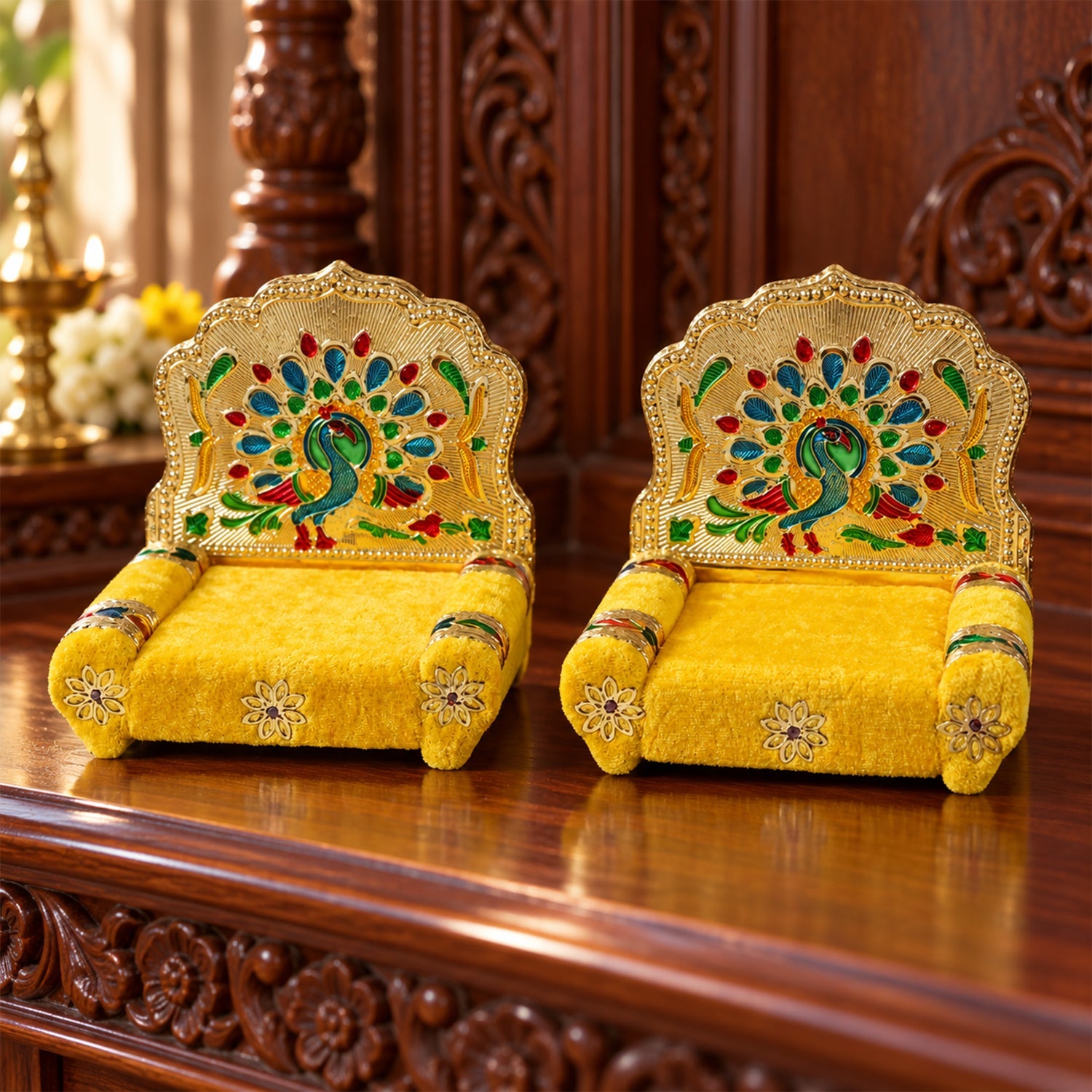 Two golden singhasans with yellow seats and vibrant peacock designs, placed on a dark wooden altar with blurred background