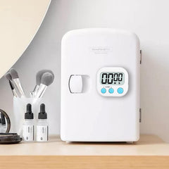 A white digital kitchen timer with blue buttons sits on a wooden desk next to a potted plant and a pen holder.