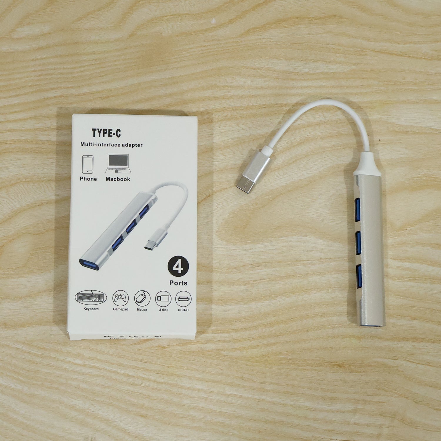A silver Type C to four-port USB hub next to its white packaging, both on a wooden surface.