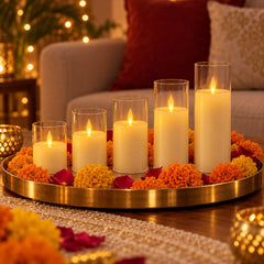Five glowing flameless LED candles in clear plastic sleeves sit on a gold tray surrounded by orange marigolds and red petals.