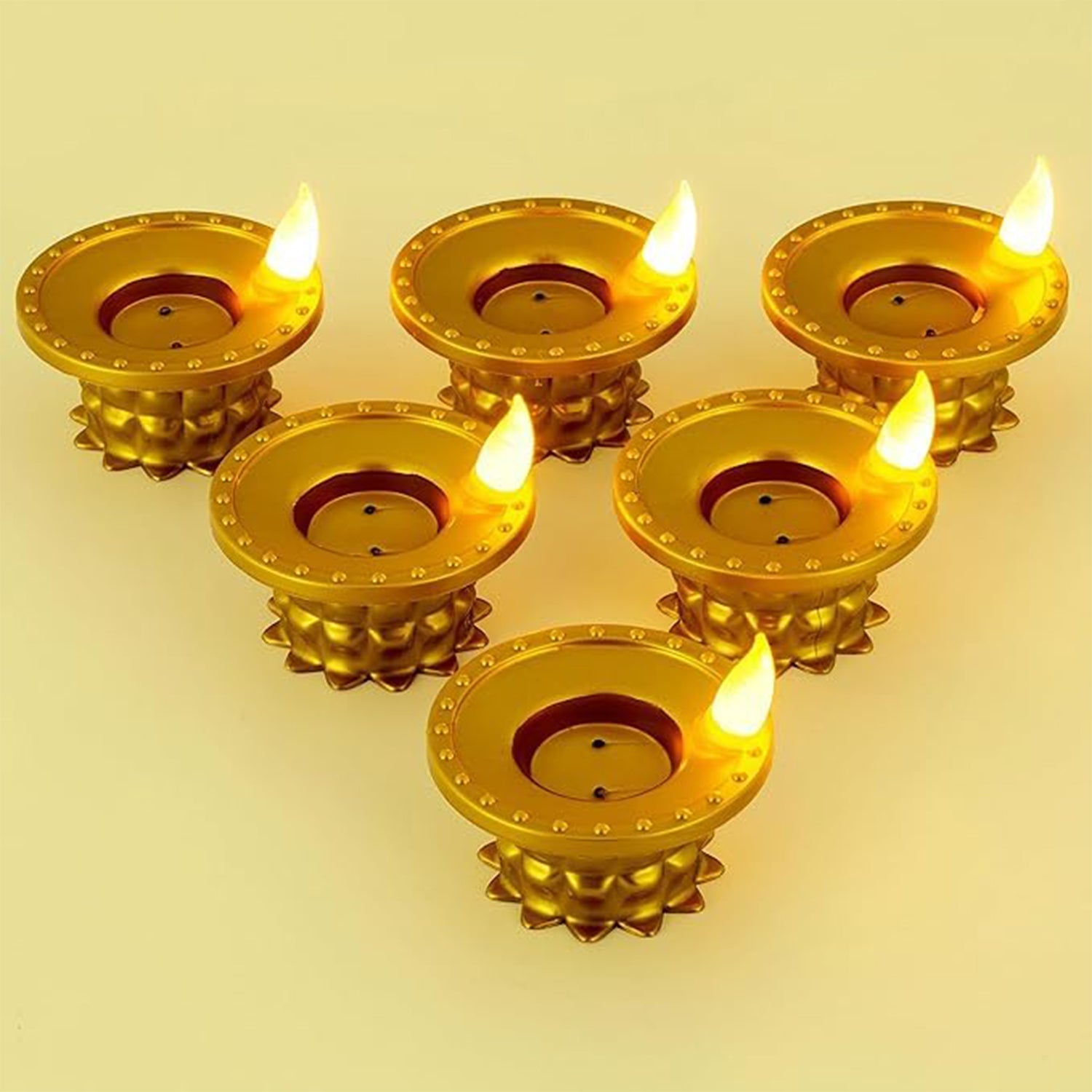 Six gold plastic LED diyas with lit flames, arranged in a staggered pattern on a light-colored surface.