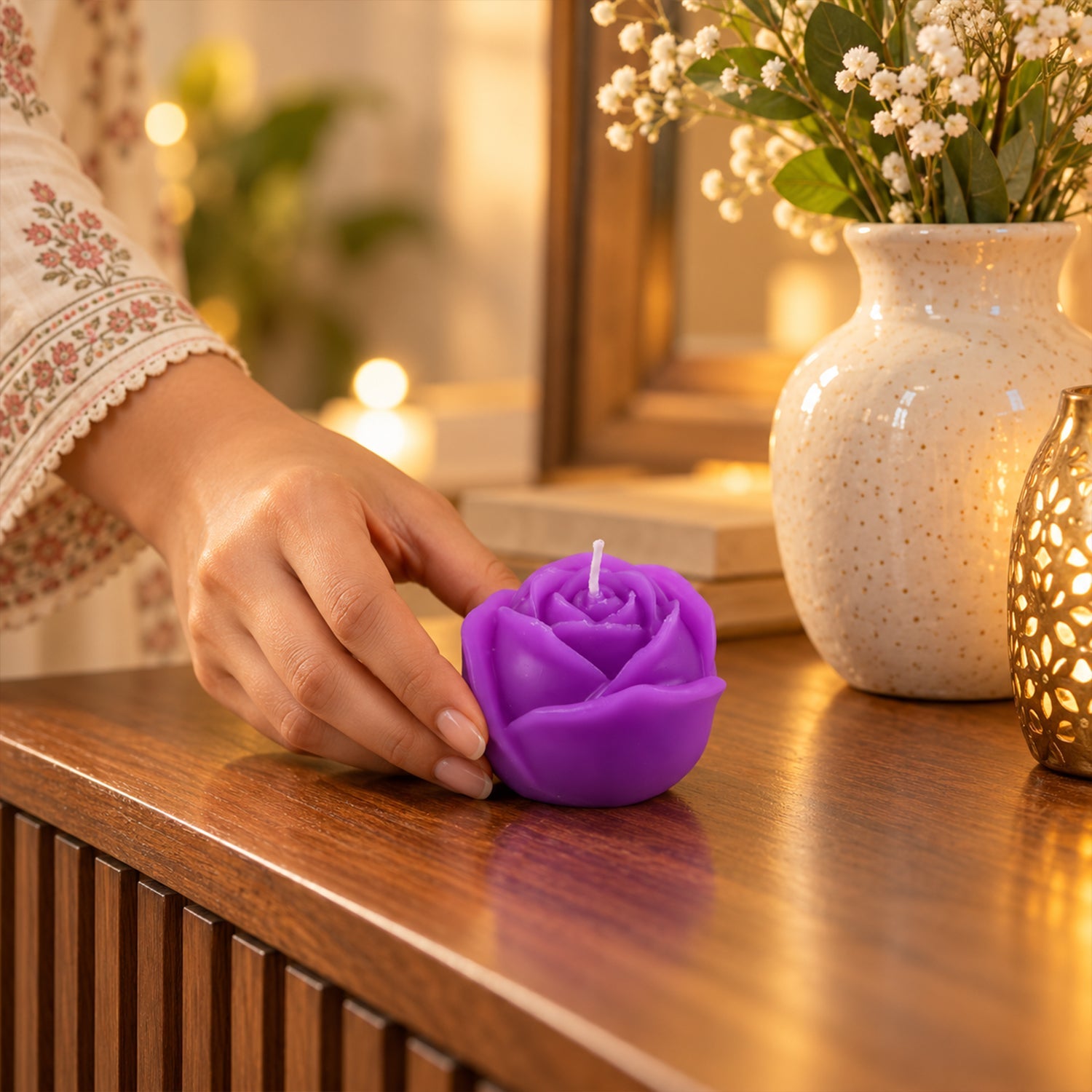 A hand gently places a purple rose-shaped candle on a ribbed wooden dresser next to a white vase with flowers.