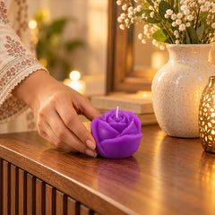 A hand gently places a purple rose-shaped candle on a ribbed wooden dresser next to a white vase with flowers.