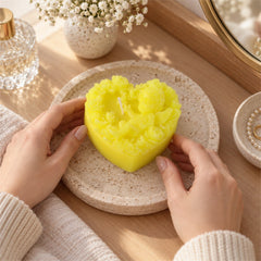 Hands gently holding a yellow heart-shaped candle with a cherub and flower design on a textured round tray.