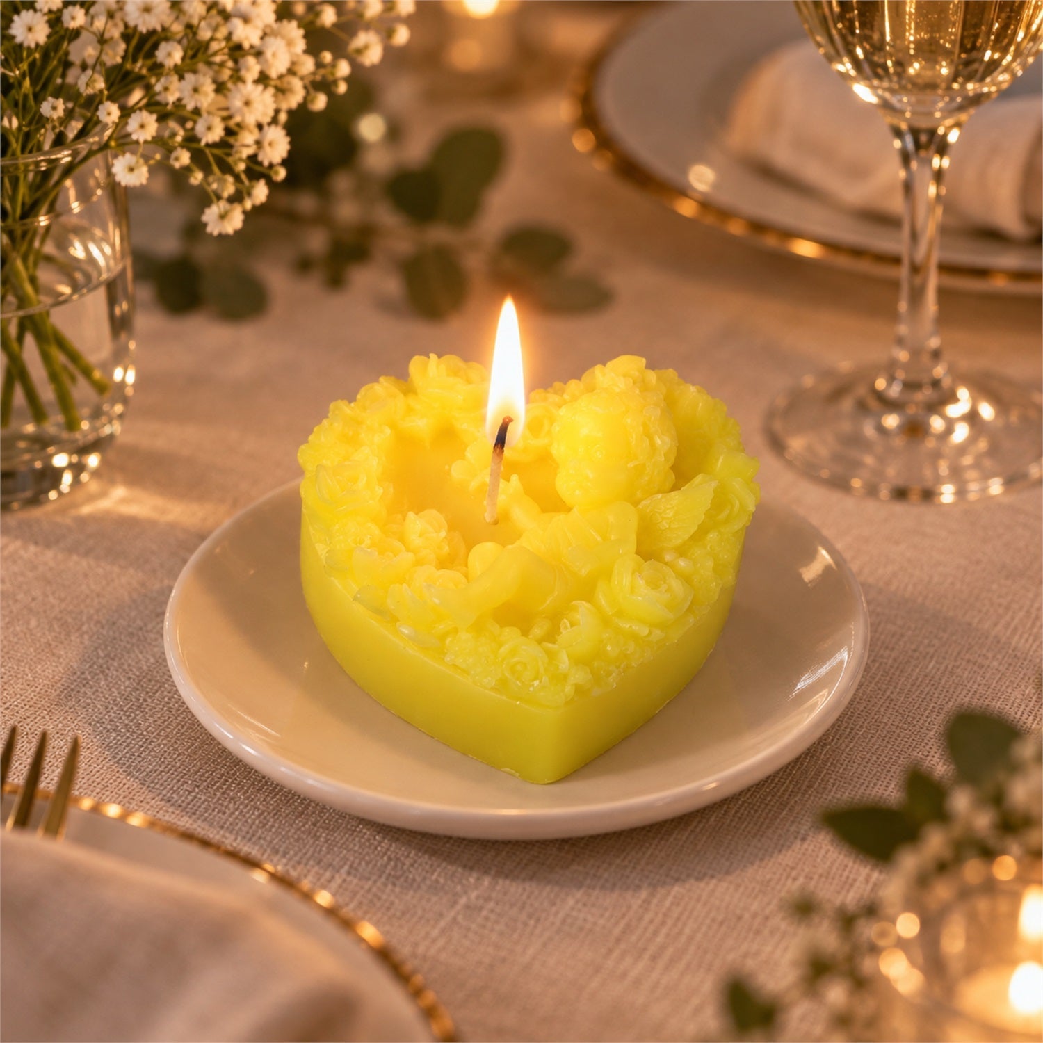 A lit yellow heart-shaped candle with a cherub and floral pattern on a white plate, set on a dining table.