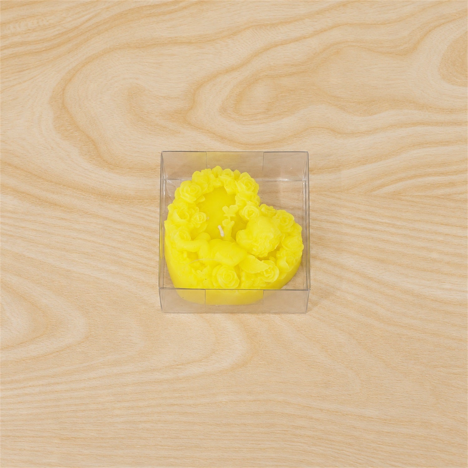 A yellow heart-shaped candle with a cherub design, neatly packaged in a clear square box on a wooden surface.