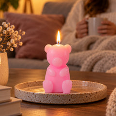 A lit pink teddy bear candle sits on a speckled ceramic plate, with a person holding a mug in the background.