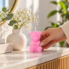 A hand gently places a pink teddy bear candle on a marble surface next to a white vase with flowers.
