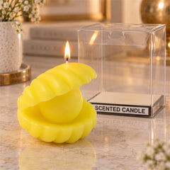 A lit yellow shell-shaped candle with a pearl center sits beside its clear plastic packaging on a marble surface.