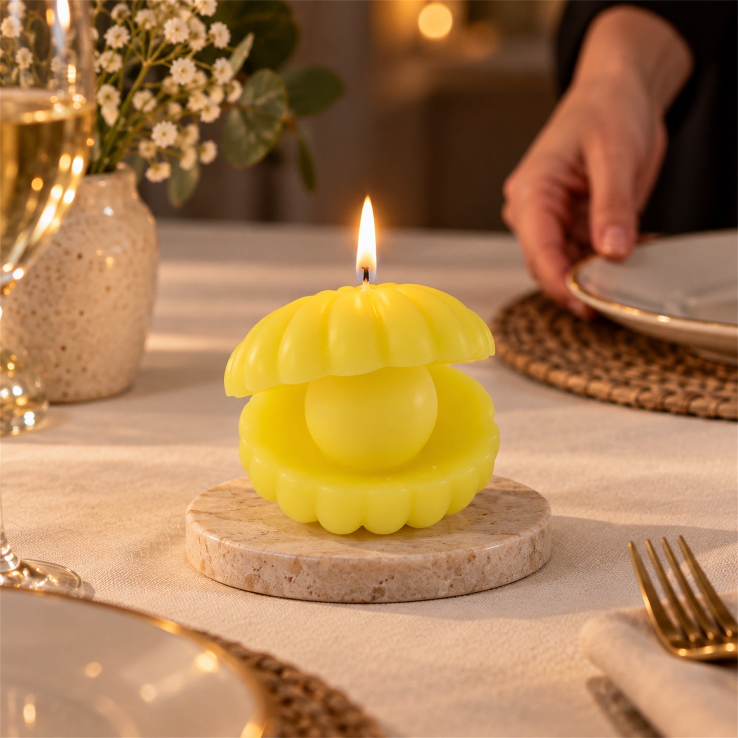 A bright yellow shell candle on a round marble coaster illuminates a dining table setting with a person in the background.