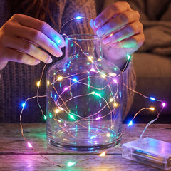 Hands gently place a string of colorful LED lights into a clear glass vase on a wooden table.