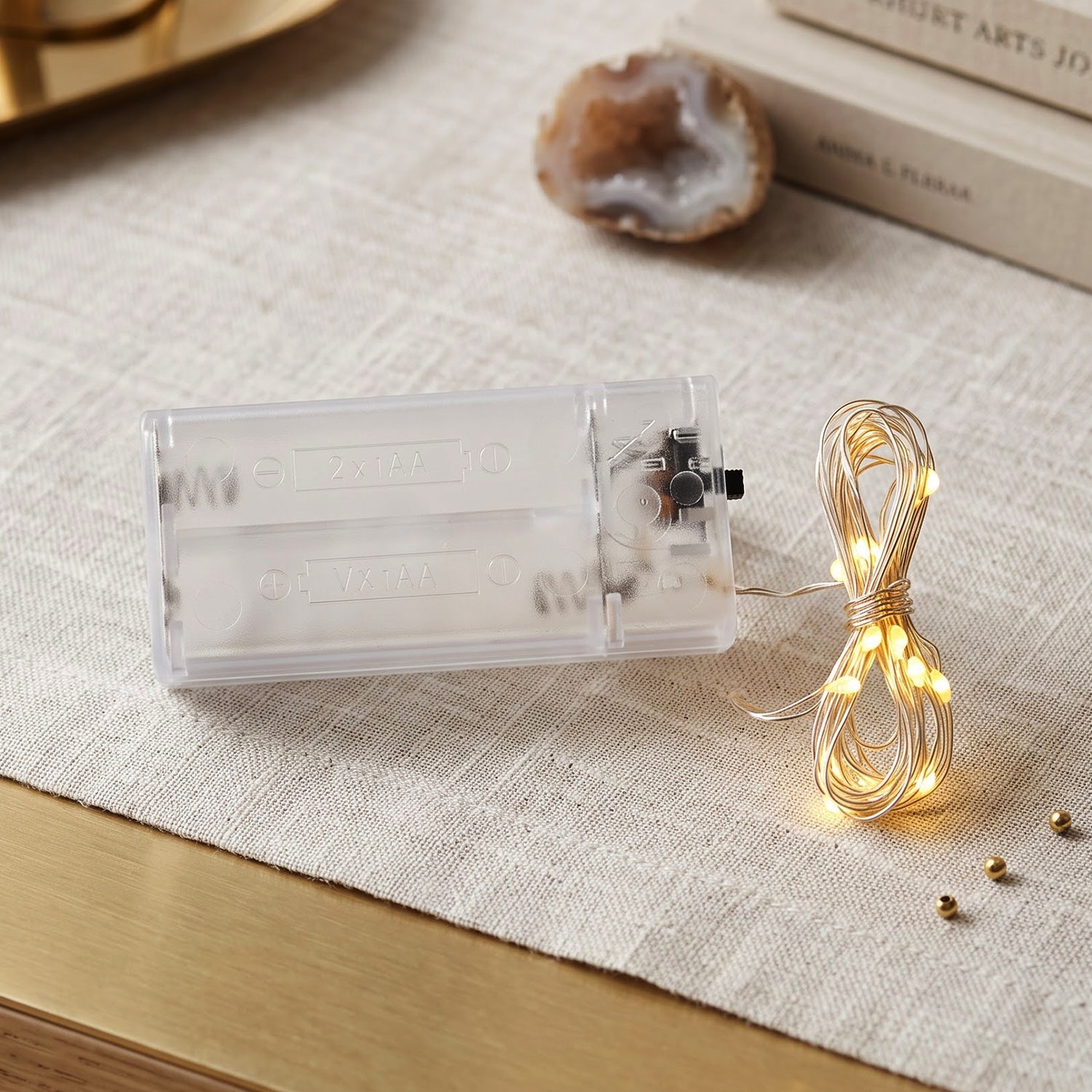 A clear battery box and coiled warm white LED string lights rest on a light linen runner with small gold beads.