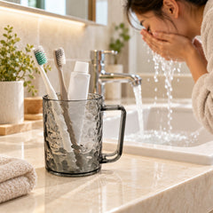 Dark grey textured plastic mug with two toothbrushes and a small white tube on a bathroom counter with a person washing