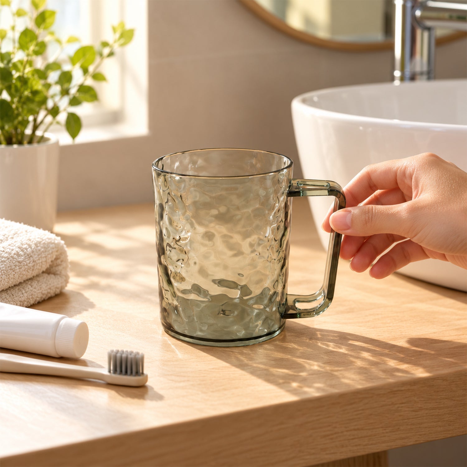 A hand reaching for a dark grey textured plastic mug on a wooden bathroom counter next to a white sink.