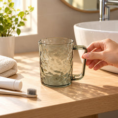 A hand reaching for a dark grey textured plastic mug on a wooden bathroom counter next to a white sink.