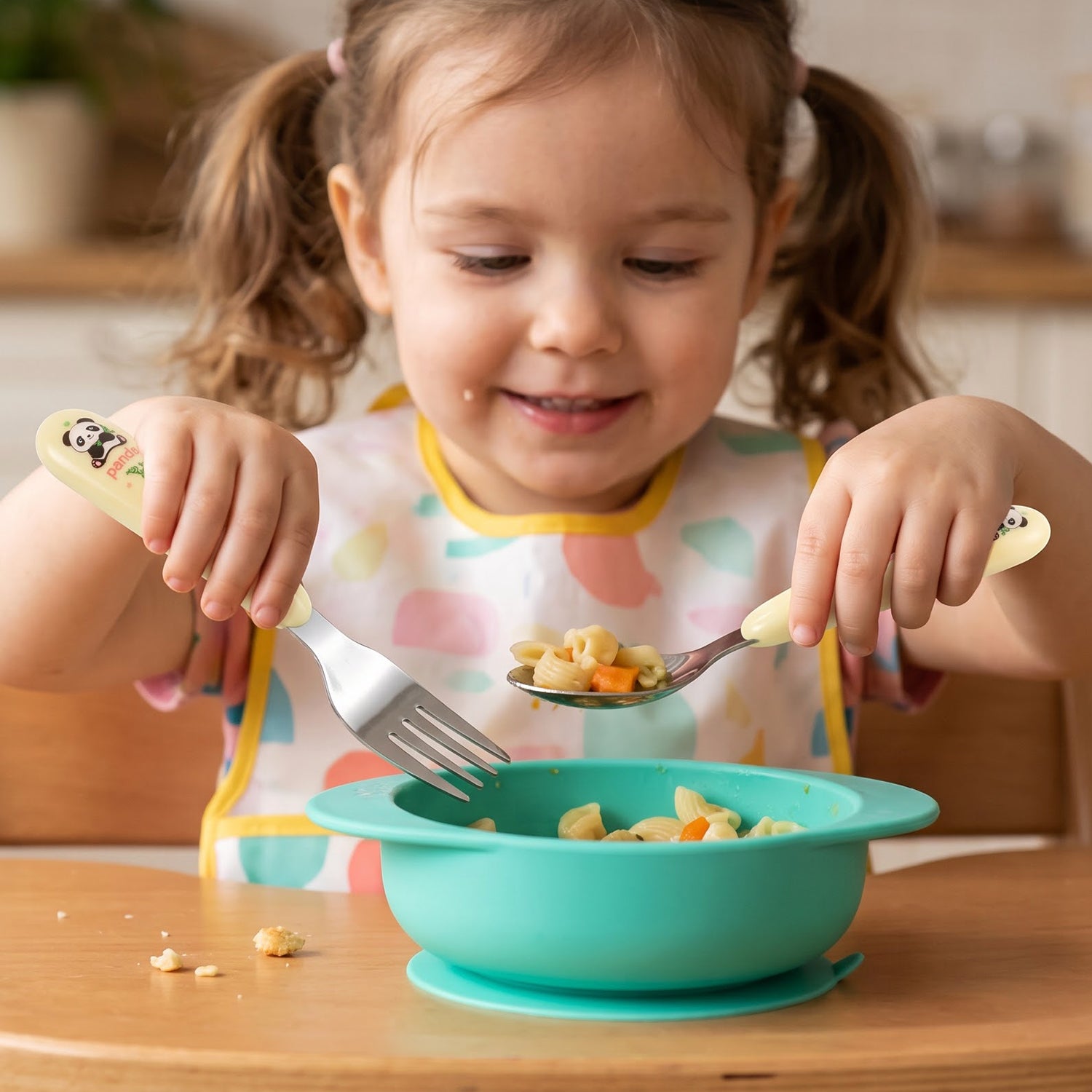 A young child with pigtails uses a yellow panda spoon and fork to eat pasta from a teal silicone bowl.