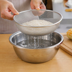 Hands hold a stainless steel mesh strainer full of white rice over a steel bowl of water.