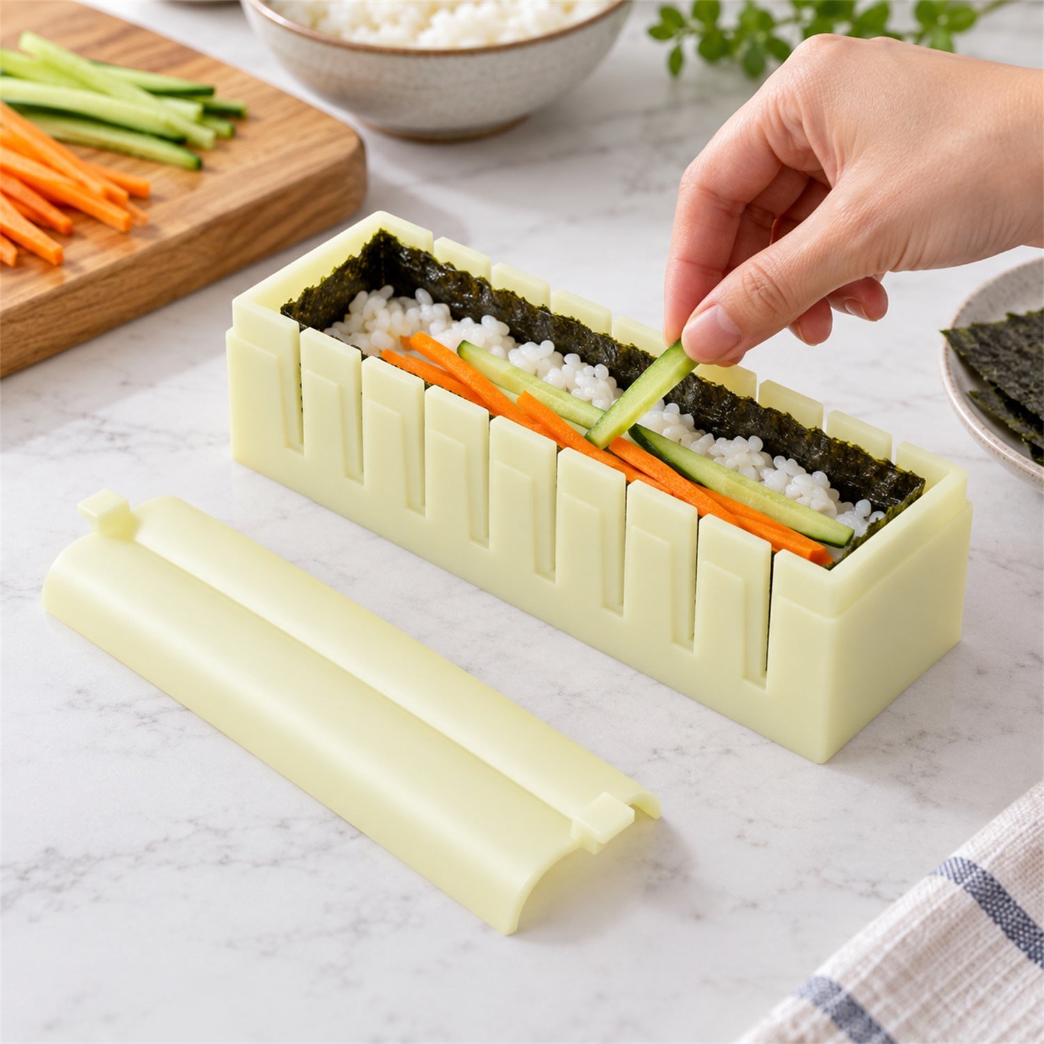 A hand places a cucumber strip into a sushi mold filled with rice and seaweed, with other ingredients visible.